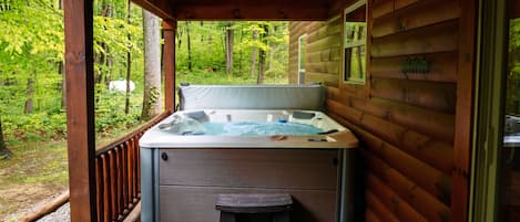 A wooden cabin with a hot tub on a deck, surrounded by a forest.
