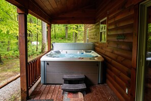A wooden cabin with a hot tub on a deck, surrounded by a forest.