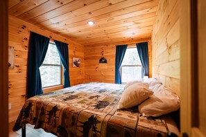 Ein Schlafzimmer einer Holzhütte mit Bett, zwei Vorhängen an Fenstern, einer Deckenleuchte und einer Wanddekoration.