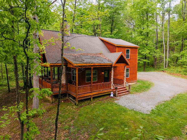 A wooden cabin with a porch, a gravel driveway, and surrounded by trees.