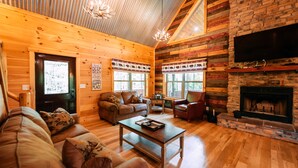 A log cabin interior with a wooden ceiling, a fireplace, a TV mounted on the wall, and a cozy seating area with a sofa and a coffee table.