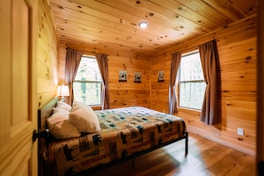 A wooden cabin room with a bed, two windows with curtains, a wall-mounted lamp, and a quilt with animal prints.