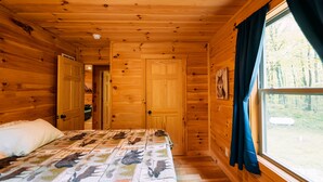 A wooden cabin bedroom with a bed, a window with curtains, and a door leading to another room. - Bear Crossing | Hocking Hills, Sleeps 10, Pets (Logan)