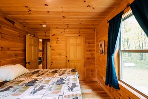 A wooden cabin bedroom with a bed, a window with curtains, and a door leading to another room.