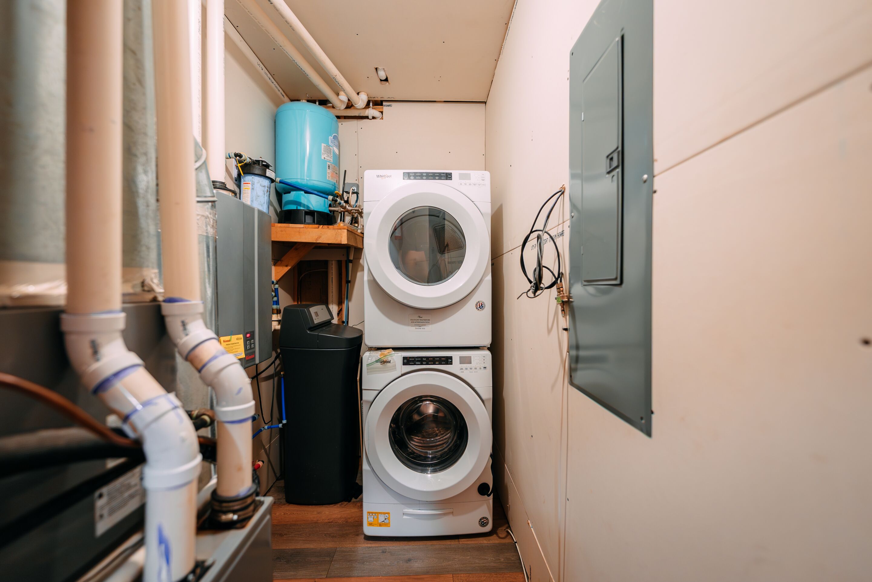 Une buanderie équipée d’un lave-linge et d’un sèche-linge superposés, d’un chauffe-eau et de tuyauteries apparentes.
