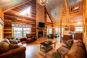 A log cabin interior with a fireplace, wooden furniture, and a loft dining area.