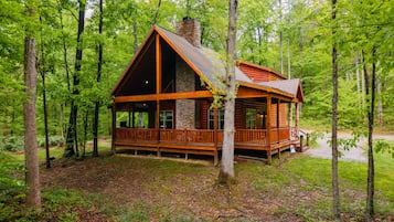 Une cabane avec une cheminée en pierre, une terrasse en bois et entourée d’arbres.