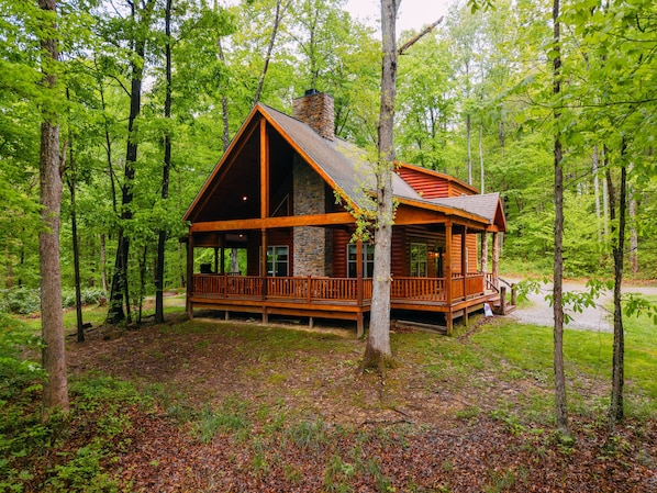 A cabin with a stone chimney, wooden deck, and surrounded by trees.