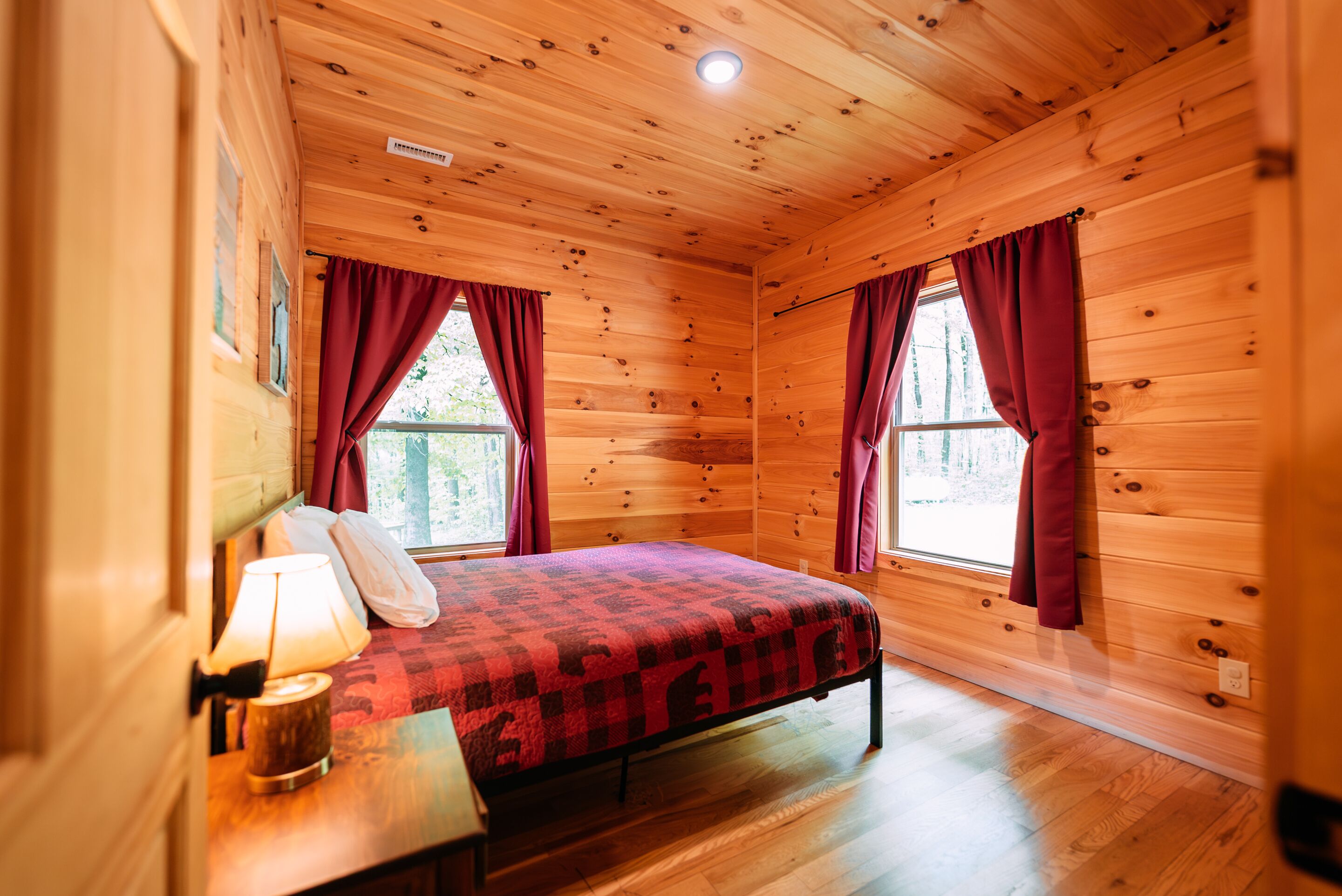 Une chambre dans un chalet en bois, avec un lit, une table de chevet, une lampe et deux fenêtres avec des rideaux.