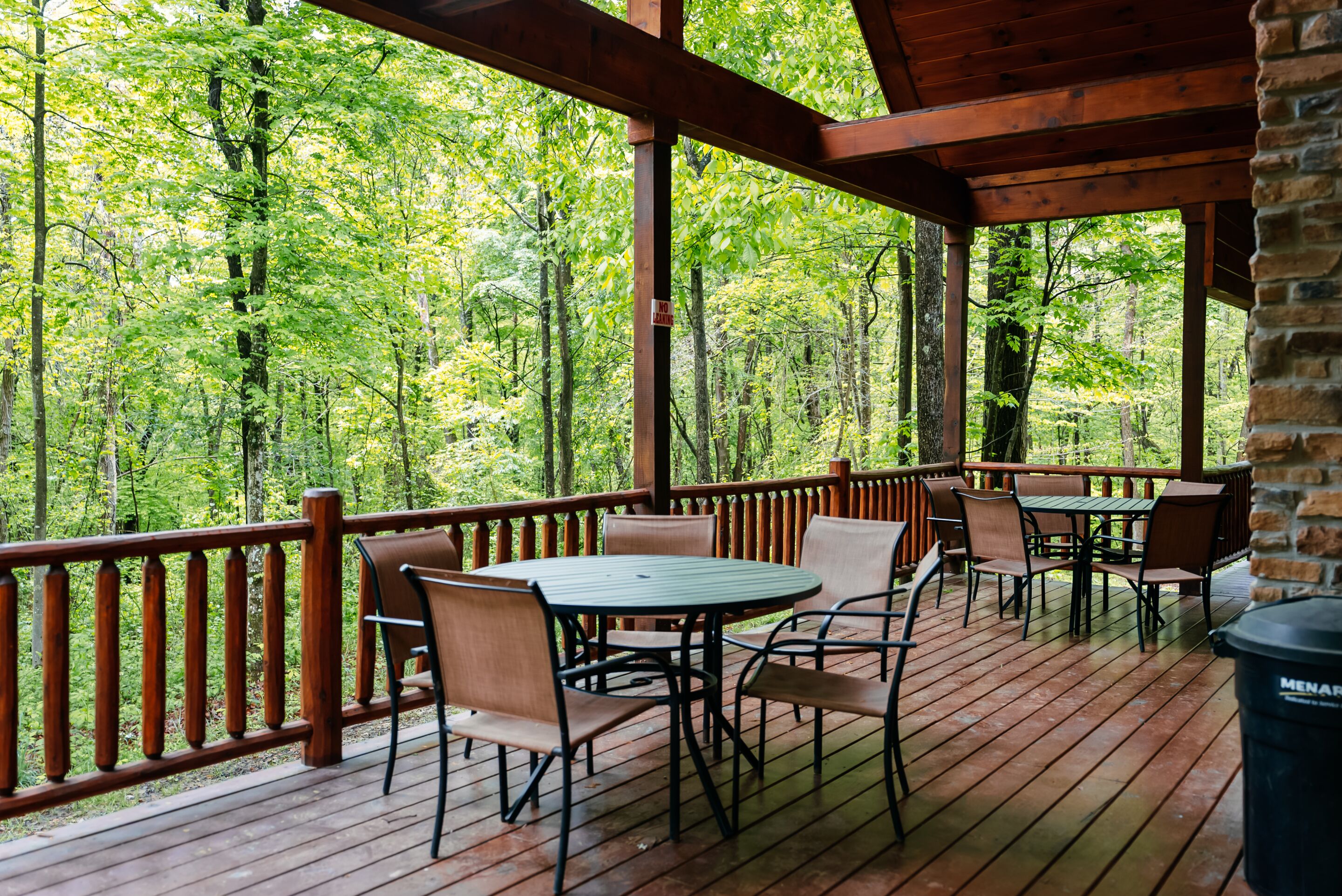 A wooden deck with tables and chairs, surrounded by a wooden railing, overlooking a forested area.
