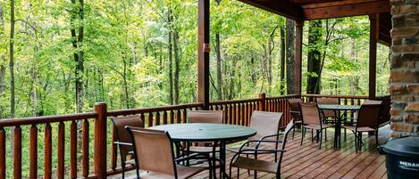 A wooden deck with tables and chairs, surrounded by a wooden railing, overlooking a forested area.