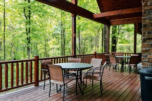 A wooden deck with tables and chairs, surrounded by a wooden railing, overlooking a forested area.