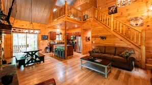 A cabin interior with a wooden staircase, a dining area, kitchen, and a living room with a sofa.