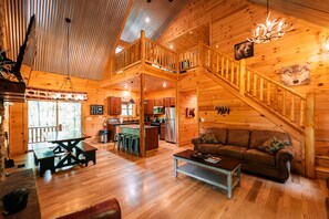 A cabin interior with a wooden staircase, a dining area, kitchen, and a living room with a sofa.
