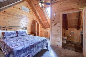 A wooden cabin bedroom with a bed, a ceiling fan, and a bathroom with a toilet and sink.
