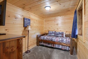 A cozy cabin room with a wooden ceiling, a bed with patterned bedding, a desk with a lamp, and a sign on the wall.