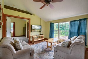 Ein Wohnzimmer mit einer Sofagarnitur, einem Couchtisch, einem Fernseher und einem großen Fenster mit Vorhängen.