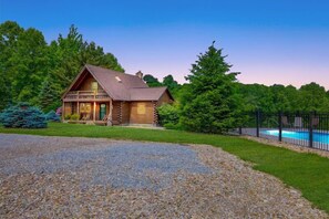 A log cabin with a porch, surrounded by a gravel driveway and a pool.