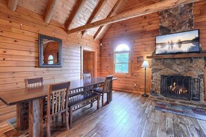 A wooden cabin interior with a dining table, chairs, a stone fireplace, and a television mounted above it.