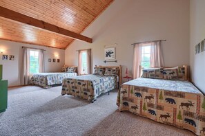 A cozy room with three beds, each with a bear-themed bedspread, wooden ceiling beams, and a large window with curtains.