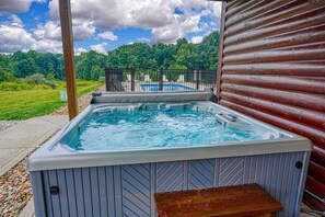 A hot tub with a wooden deck and a view of a swimming pool and forest.