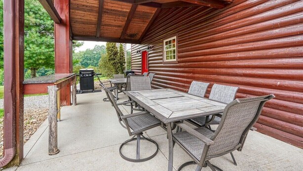 Eine überdachte Terrasse mit einem langen Esstisch und Stühlen, einer roten Tür und Blick auf eine bewaldete Gegend.
