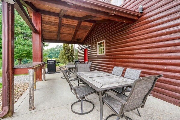 A covered outdoor patio with a long dining table and chairs, a red door, and a view of a forested area.