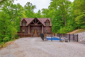 A log cabin with a front porch, a swimming pool, and a gravel driveway.