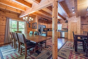 A wooden interior with a dining table, chairs, a chandelier, and a kitchen area.