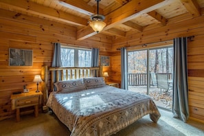 A log cabin bedroom with a bed, two bedside tables, a ceiling fan, and large windows with curtains.