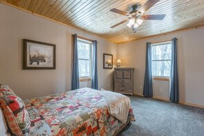 Un dormitorio con cama, ventilador de techo, cómoda y dos ventanas con cortinas.