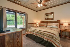 Une chambre à coucher avec un grand lit, une tête de lit en bois, deux tables de chevet avec des lampes, un ventilateur de plafond et une fenêtre avec des rideaux.