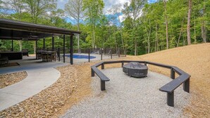 A covered outdoor seating area with tables and benches, a fire pit, and a pool in the background. - Coal Mine | Hocking Hills, 8BR, Pool & Bowling (Logan)