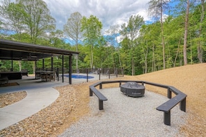 A covered outdoor seating area with tables and benches, a fire pit, and a pool in the background. - Coal Mine | Hocking Hills, 8BR, Pool & Bowling (Logan)