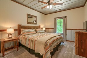 A bedroom with a bed, a ceiling fan, a mirror, a window, and a nightstand with a lamp.