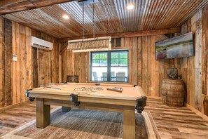 A pool table in a wooden room with a large window, a mounted TV, and an air conditioning unit.
