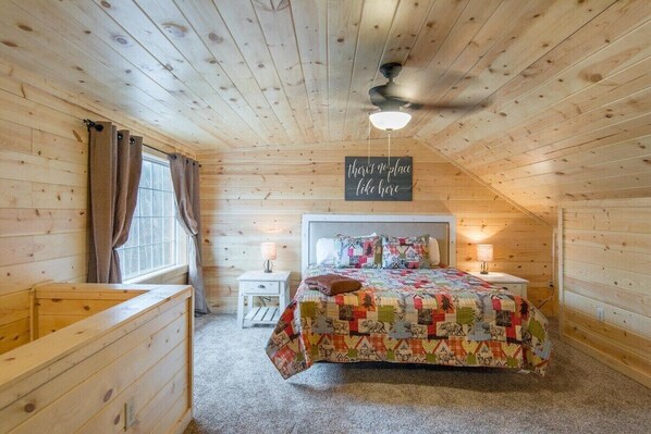 A cozy bedroom with a wooden ceiling, a bed with a colorful quilt, a nightstand, and a sign above the bed.