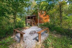 A rustic cabin with a wooden deck and a fire pit in a forest setting.