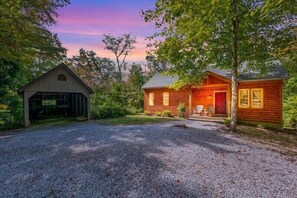 A rustic cabin with a gravel driveway, surrounded by trees and a wooden barn.