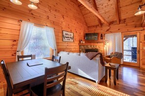 A cozy log cabin interior with a wooden dining table, a sofa, a bed, and a fireplace.
