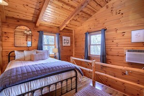 A wooden cabin bedroom with a bed, a mirror, two windows with curtains, and a wall-mounted air conditioner. - Tranquility Ridge | Hocking Hills, 5 Acres, Pets (Logan)