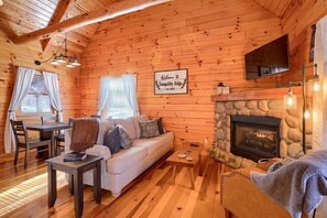 A cozy log cabin interior with a stone fireplace, wooden walls, a sofa, and a dining area. - Tranquility Ridge | Hocking Hills, 5 Acres, Pets (Logan)
