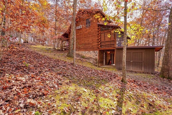 A rustic log cabin with a stone foundation, a wooden porch, and a small shed.