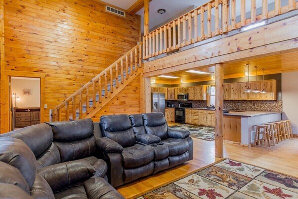 A spacious living area with a black leather sofa, wooden flooring, and a kitchen in the background.