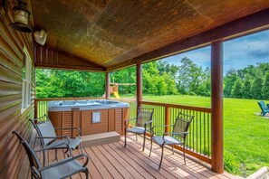 A wooden deck with a hot tub, chairs, and a view of a grassy area with trees. - Dragonfly Lodge | Hocking Hills, 5BR, Hot Tub (Logan)