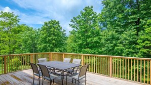 A wooden deck with a table and chairs, surrounded by a wooden railing, overlooking a dense forest.