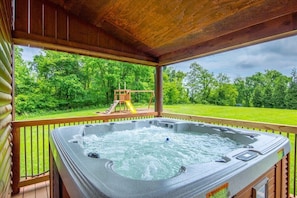 A hot tub on a wooden deck with a view of a playground and lush greenery. - Dragonfly Lodge | Hocking Hills, 5BR, Hot Tub (Logan)