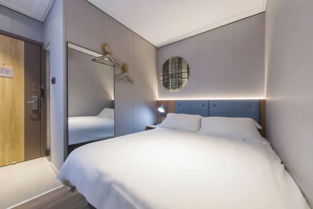 A modern hotel room with a large bed, a mirror, a wall-mounted light, and a wooden door.