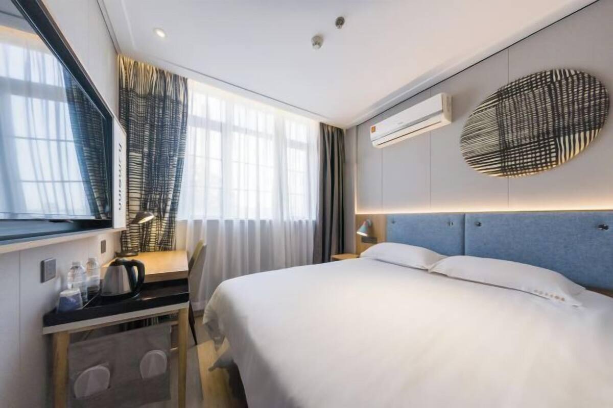 A modern hotel room with a bed, bedside table, lamp, air conditioning unit, and a window with curtains.
