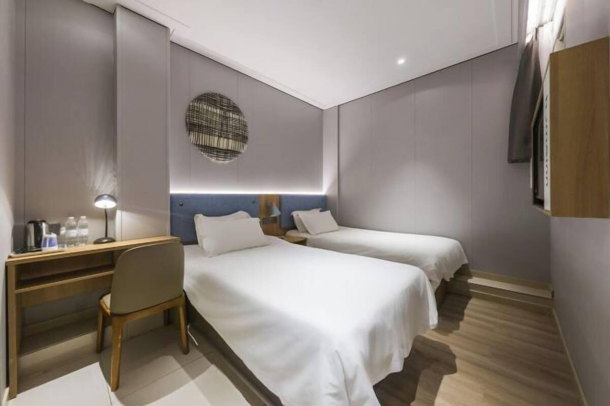 A modern hotel room with two single beds, a desk, a chair, and a wall-mounted TV.